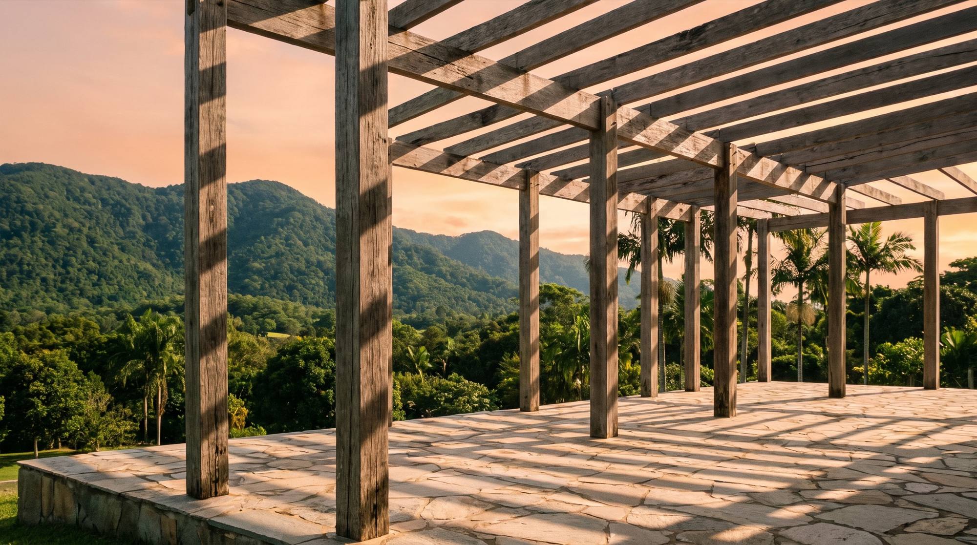A timber pergola frame over a paved terrace with rainforest hills beyond at sunset