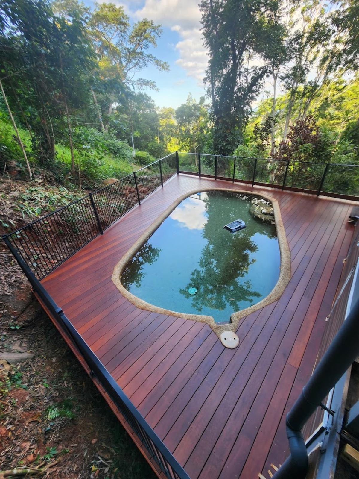 A hardwood deck wrapping a swimming pool, with black fencing and rainforest behind
